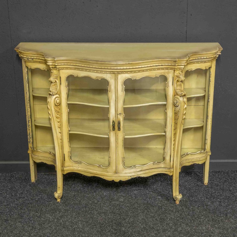 An antique cream painted serpentine credenza featuring central glazed doors, carved scroll accents, and open side shelves.