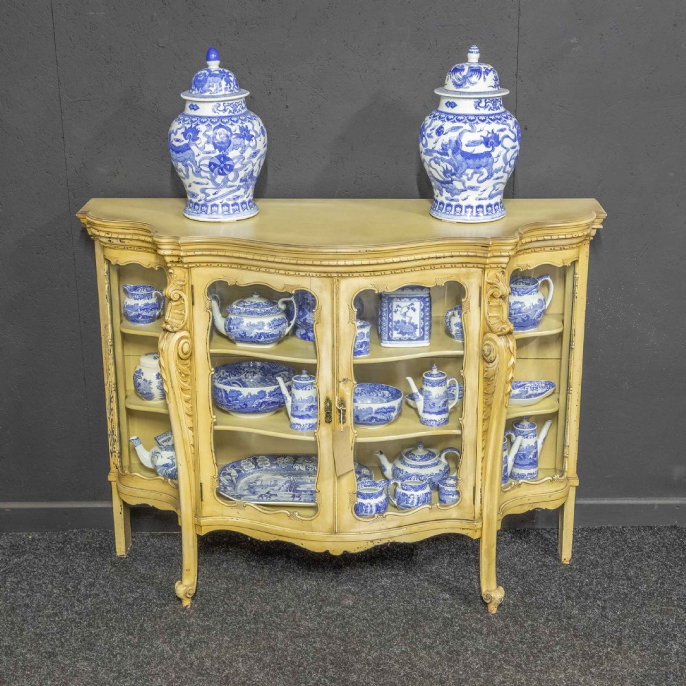 An antique cream painted serpentine credenza featuring central glazed doors, carved scroll accents, and open side shelves. View II