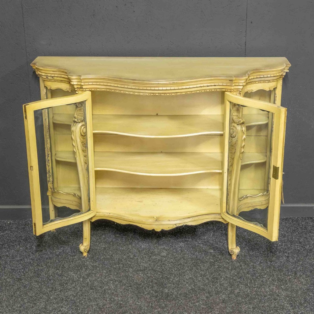An antique cream painted serpentine credenza featuring central glazed doors, carved scroll accents, and open side shelves. View III