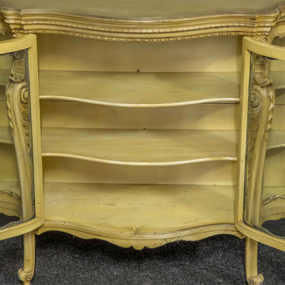 An antique cream painted serpentine credenza featuring central glazed doors, carved scroll accents, and open side shelves. View IV
