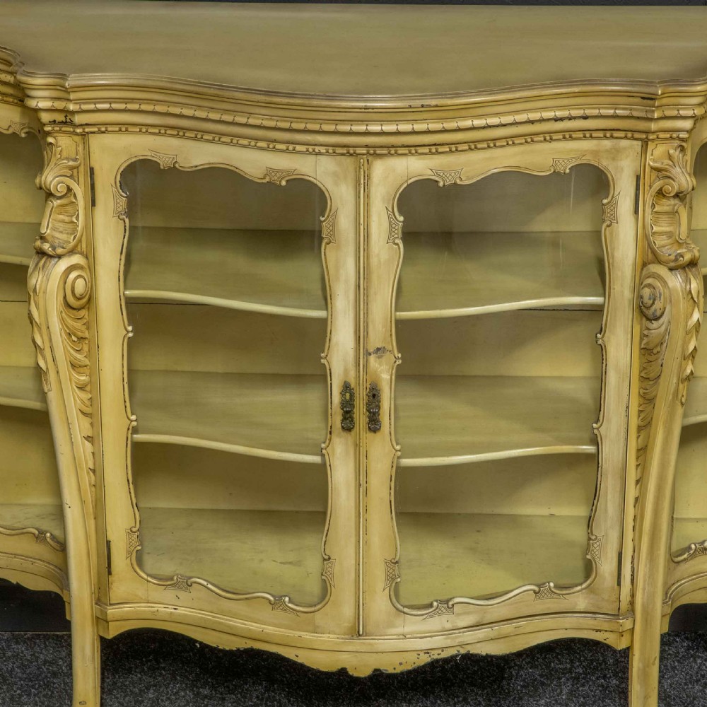 An antique cream painted serpentine credenza featuring central glazed doors, carved scroll accents, and open side shelves. View V