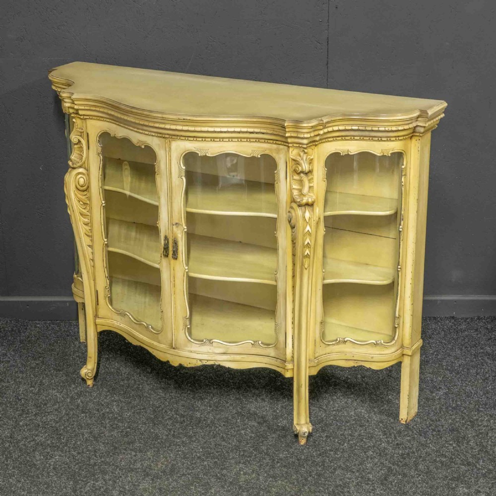 An antique cream painted serpentine credenza featuring central glazed doors, carved scroll accents, and open side shelves. View VI