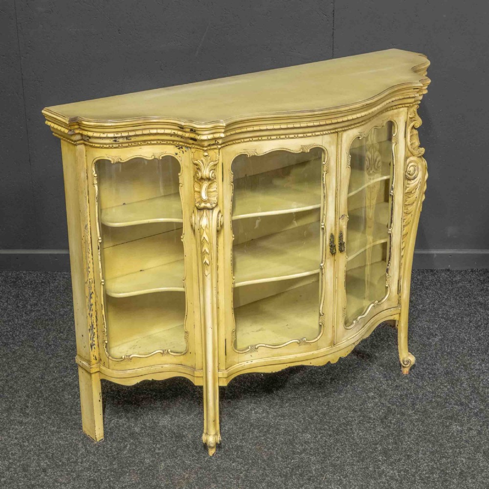An antique cream painted serpentine credenza featuring central glazed doors, carved scroll accents, and open side shelves. View VIII