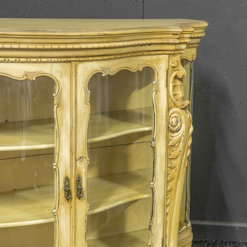 An antique cream painted serpentine credenza featuring central glazed doors, carved scroll accents, and open side shelves. View IX