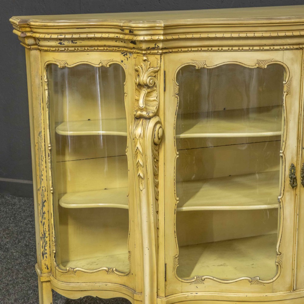 An antique cream painted serpentine credenza featuring central glazed doors, carved scroll accents, and open side shelves. View X