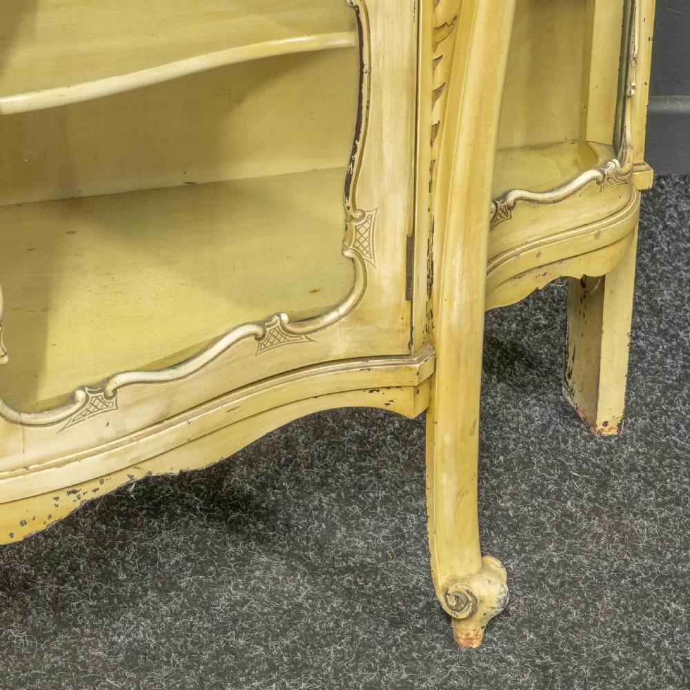 An antique cream painted serpentine credenza featuring central glazed doors, carved scroll accents, and open side shelves. View XI
