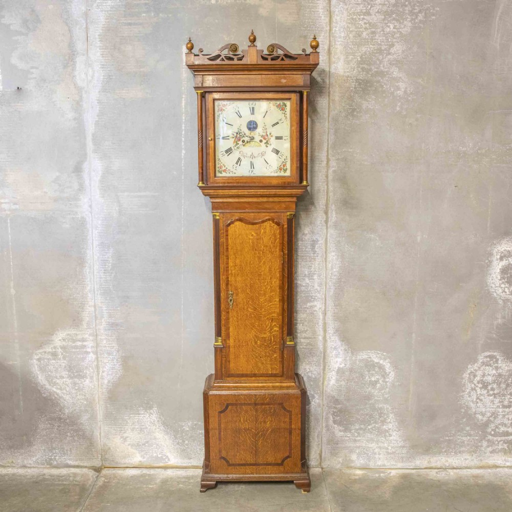 An antique Georgian oak longcase clock featuring a carved fretted pediment, brass finials, and a hand-painted square dial face.