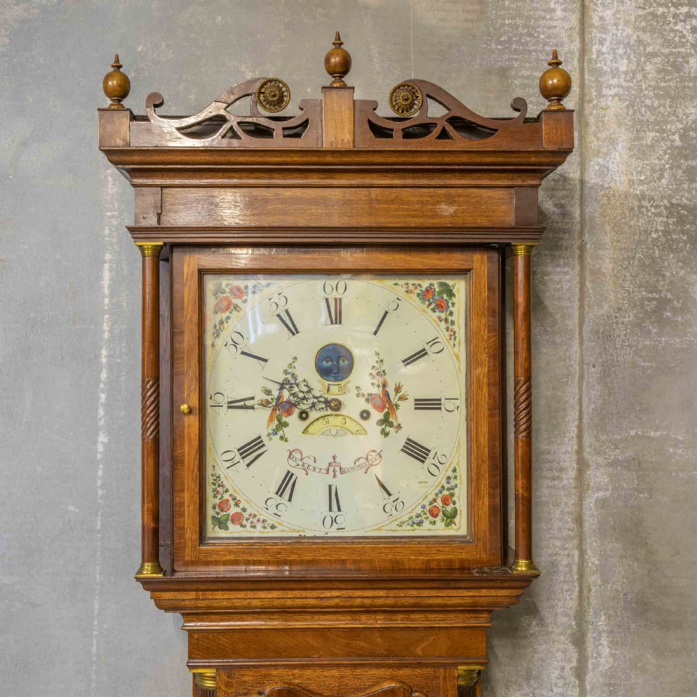 An antique Georgian oak longcase clock featuring a carved fretted pediment, brass finials, and a hand-painted square dial face. View V