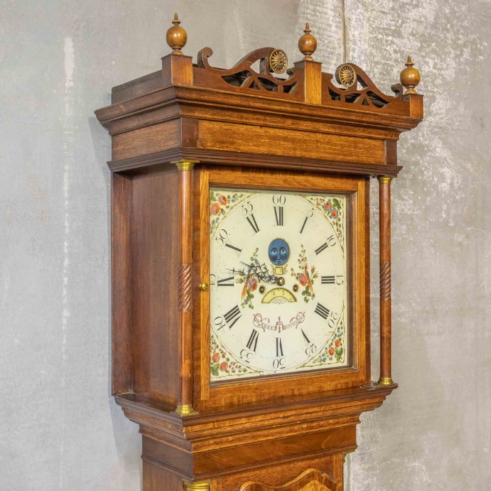 An antique Georgian oak longcase clock featuring a carved fretted pediment, brass finials, and a hand-painted square dial face. View X