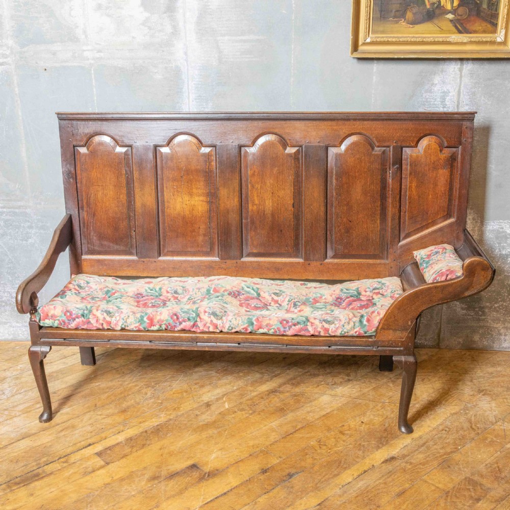 An antique dark oak sleeper settle featuring a high panelled backrest, cabriole front legs, and a floral upholstered cushion.