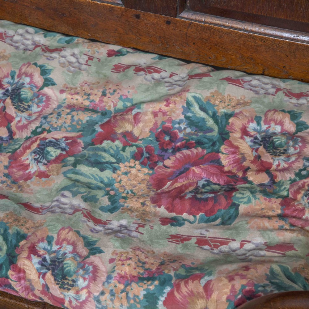 An antique dark oak sleeper settle featuring a high panelled backrest, cabriole front legs, and a floral upholstered cushion. View X