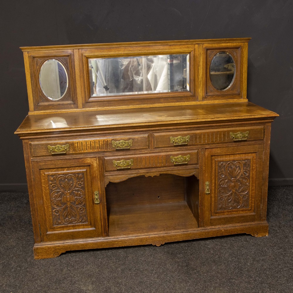 An Edwardian carved oak sideboard featuring a mirrored back gallery, relief-carved door panels, multiple drawers with brass handles, and a central open display shelf.