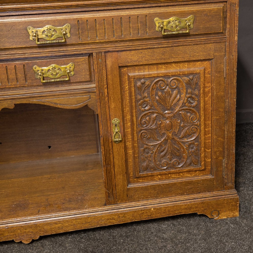 An Edwardian carved oak sideboard featuring a mirrored back gallery, relief-carved door panels, multiple drawers with brass handles, and a central open display shelf. View IV