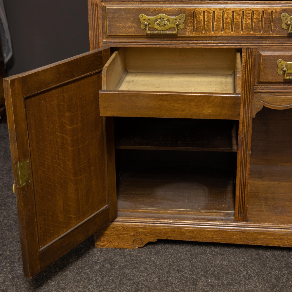 An Edwardian carved oak sideboard featuring a mirrored back gallery, relief-carved door panels, multiple drawers with brass handles, and a central open display shelf. View V