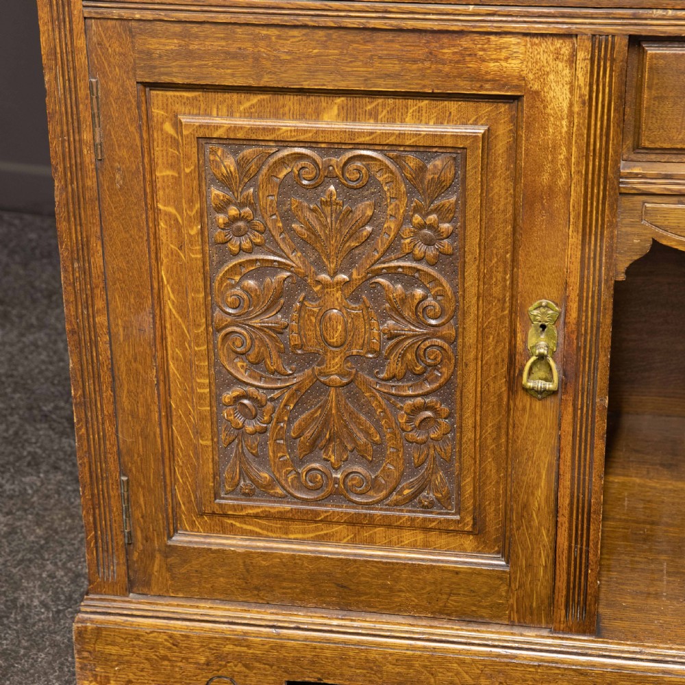 An Edwardian carved oak sideboard featuring a mirrored back gallery, relief-carved door panels, multiple drawers with brass handles, and a central open display shelf. View VI