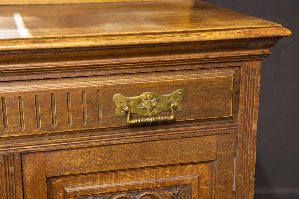 An Edwardian carved oak sideboard featuring a mirrored back gallery, relief-carved door panels, multiple drawers with brass handles, and a central open display shelf. View VII