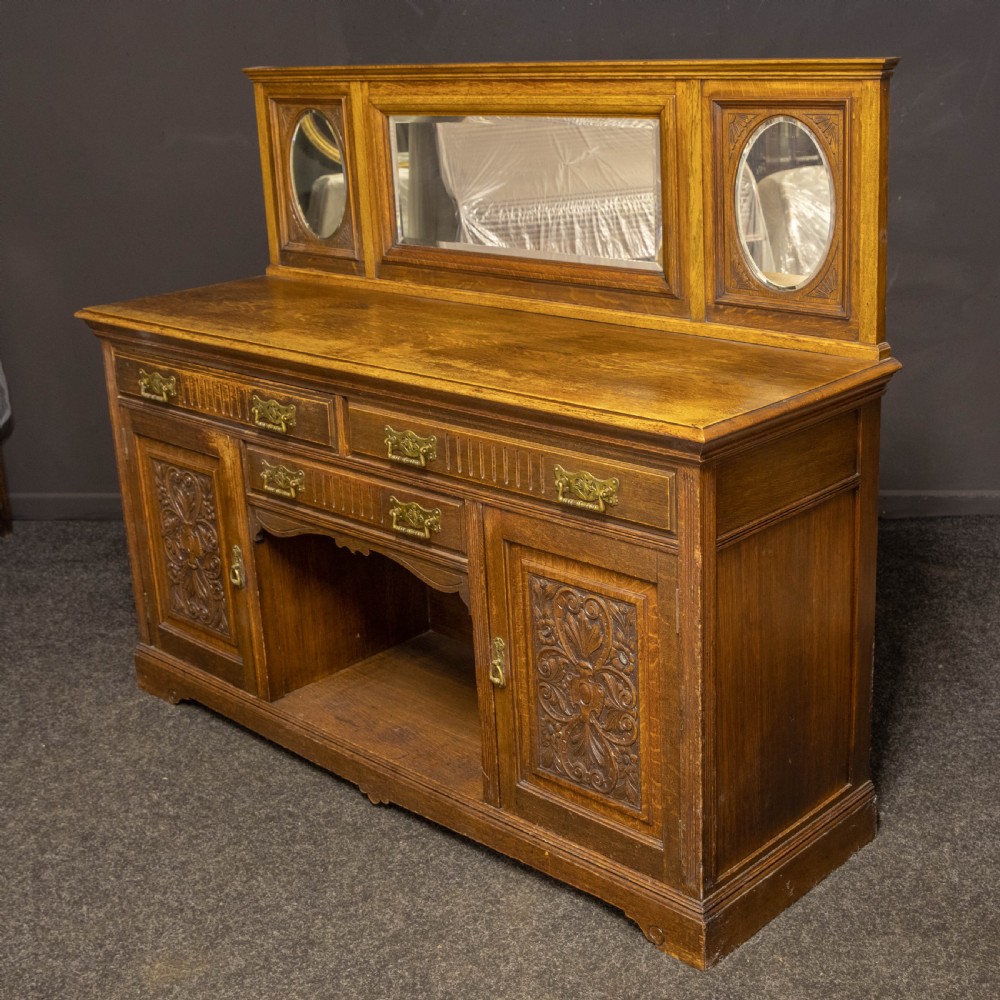 An Edwardian carved oak sideboard featuring a mirrored back gallery, relief-carved door panels, multiple drawers with brass handles, and a central open display shelf. View VIII