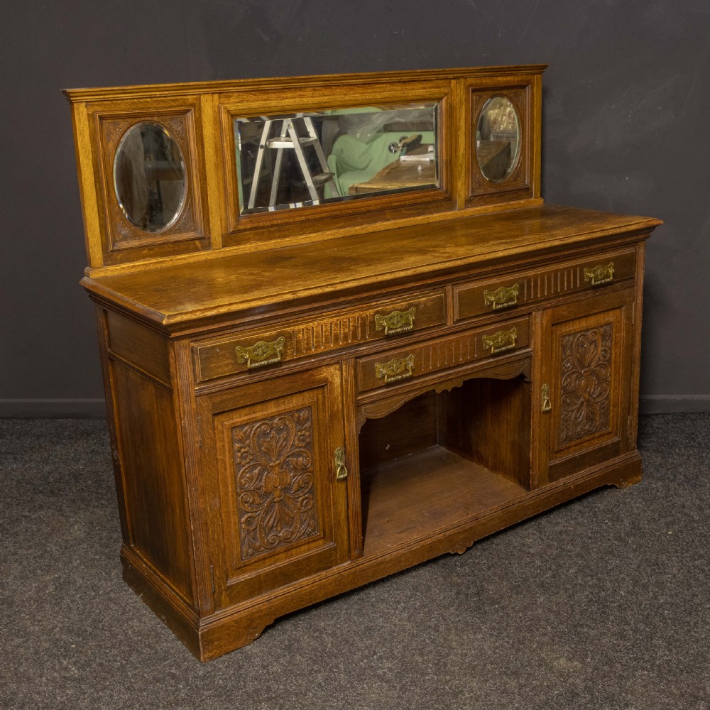 An Edwardian carved oak sideboard featuring a mirrored back gallery, relief-carved door panels, multiple drawers with brass handles, and a central open display shelf. View IX