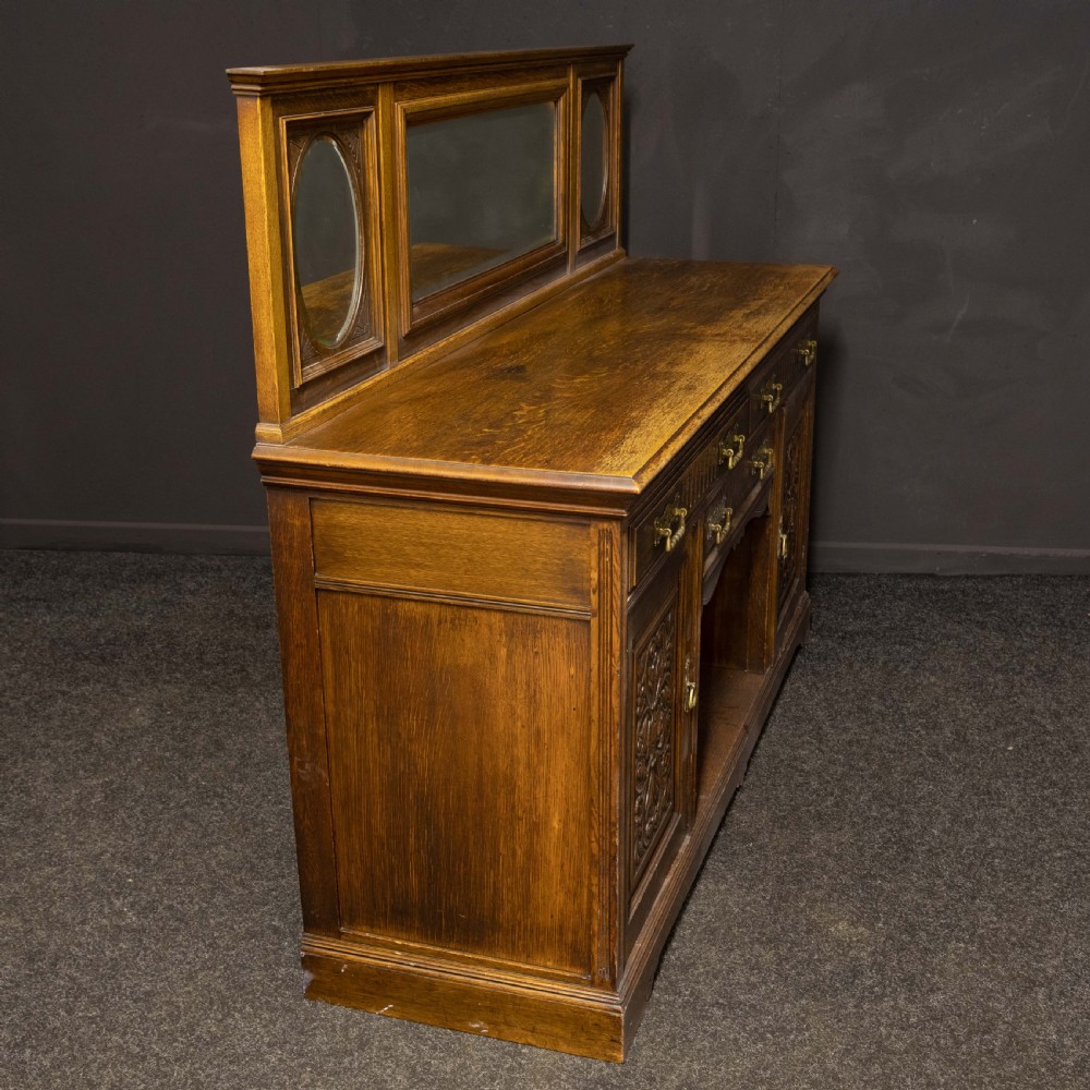 An Edwardian carved oak sideboard featuring a mirrored back gallery, relief-carved door panels, multiple drawers with brass handles, and a central open display shelf. View X