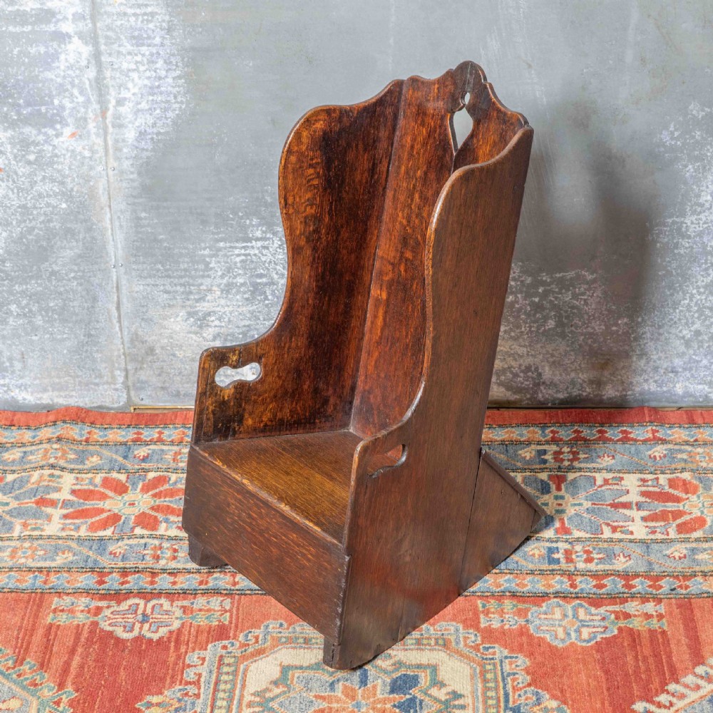 A small antique dark oak childs lambing chair featuring shaped wing sides, an enclosed back, cut-out carrying handles, and a solid box seat base.