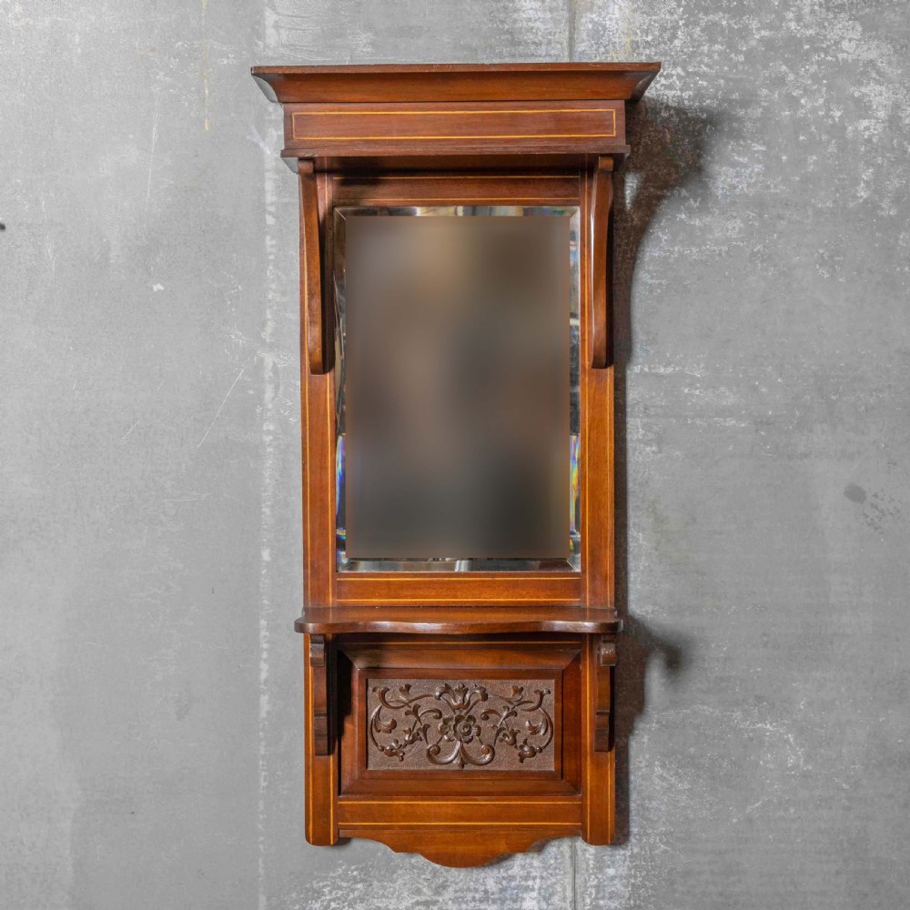An Edwardian wall-mounted mahogany mirror featuring a top shelf cornice, a central bevelled glass panel, and a carved foliate wooden panel at the base.