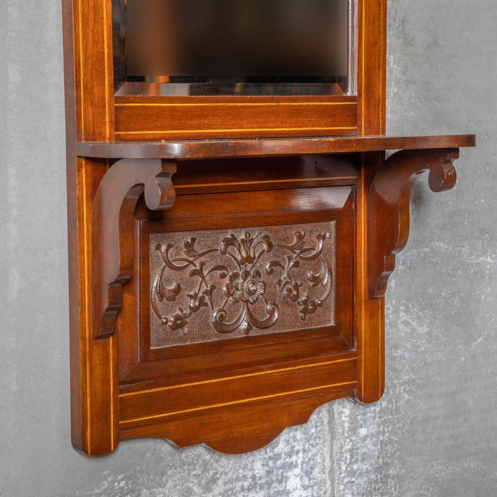 An Edwardian wall-mounted mahogany mirror featuring a top shelf cornice, a central bevelled glass panel, and a carved foliate wooden panel at the base. View II