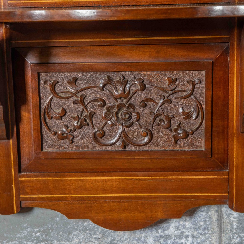 An Edwardian wall-mounted mahogany mirror featuring a top shelf cornice, a central bevelled glass panel, and a carved foliate wooden panel at the base. View III