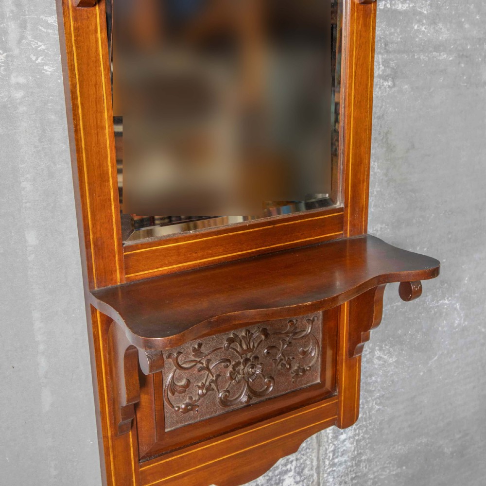 An Edwardian wall-mounted mahogany mirror featuring a top shelf cornice, a central bevelled glass panel, and a carved foliate wooden panel at the base. View IV