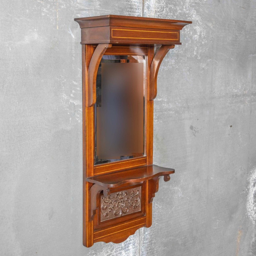 An Edwardian wall-mounted mahogany mirror featuring a top shelf cornice, a central bevelled glass panel, and a carved foliate wooden panel at the base. View VI
