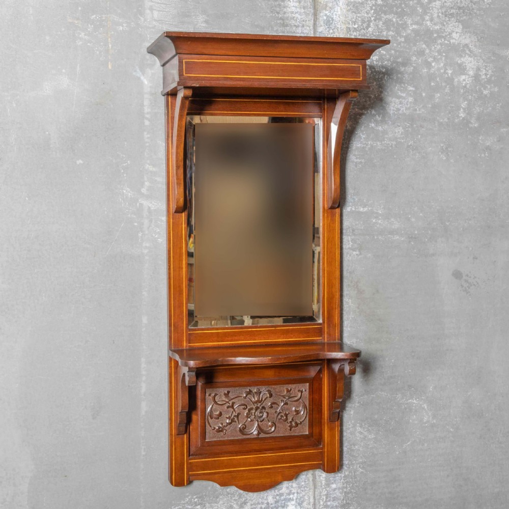 An Edwardian wall-mounted mahogany mirror featuring a top shelf cornice, a central bevelled glass panel, and a carved foliate wooden panel at the base. View VII