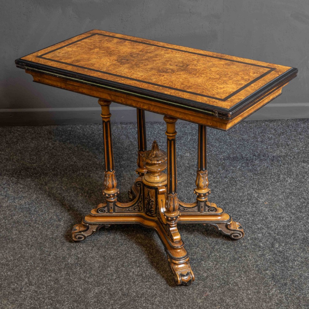 A Victorian Aesthetic Movement card table featuring burr walnut veneer, ebonised borders, turned support columns, and a central carved finial.