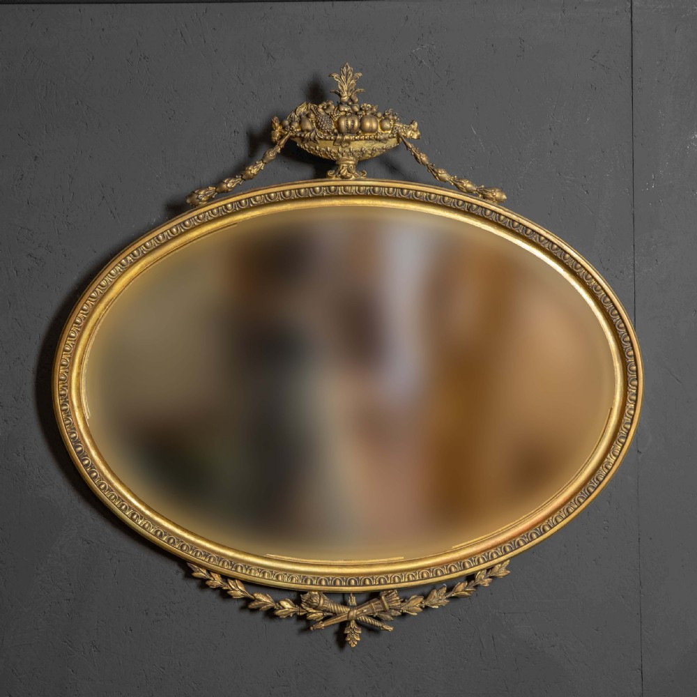 An oval gilded wall mirror decorated with an ornamental urn cresting at the top and floral swags along the lower edge.