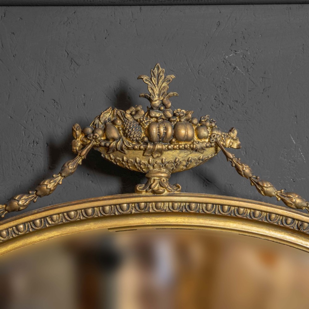 An oval gilded wall mirror decorated with an ornamental urn cresting at the top and floral swags along the lower edge. View II