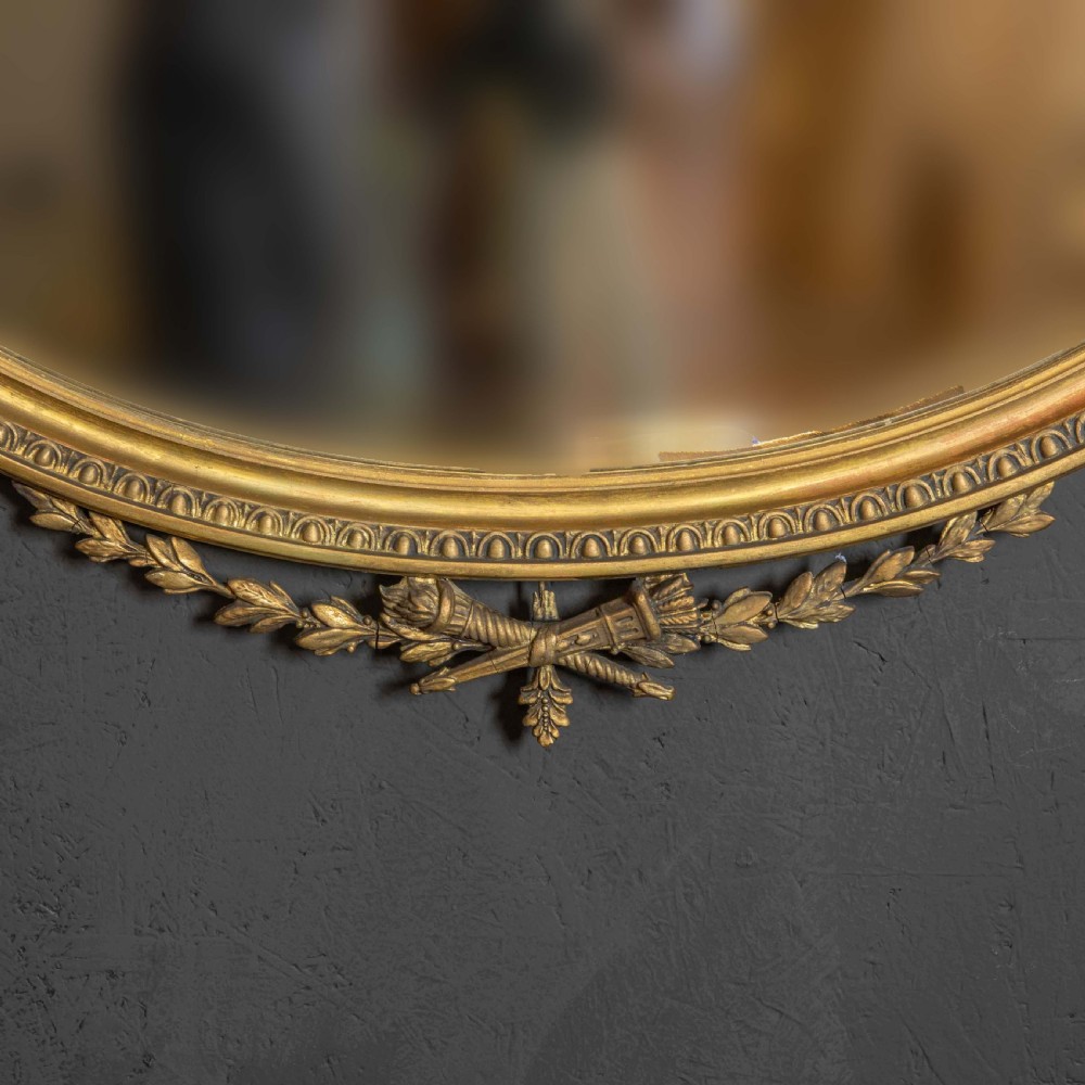 An oval gilded wall mirror decorated with an ornamental urn cresting at the top and floral swags along the lower edge. View IV
