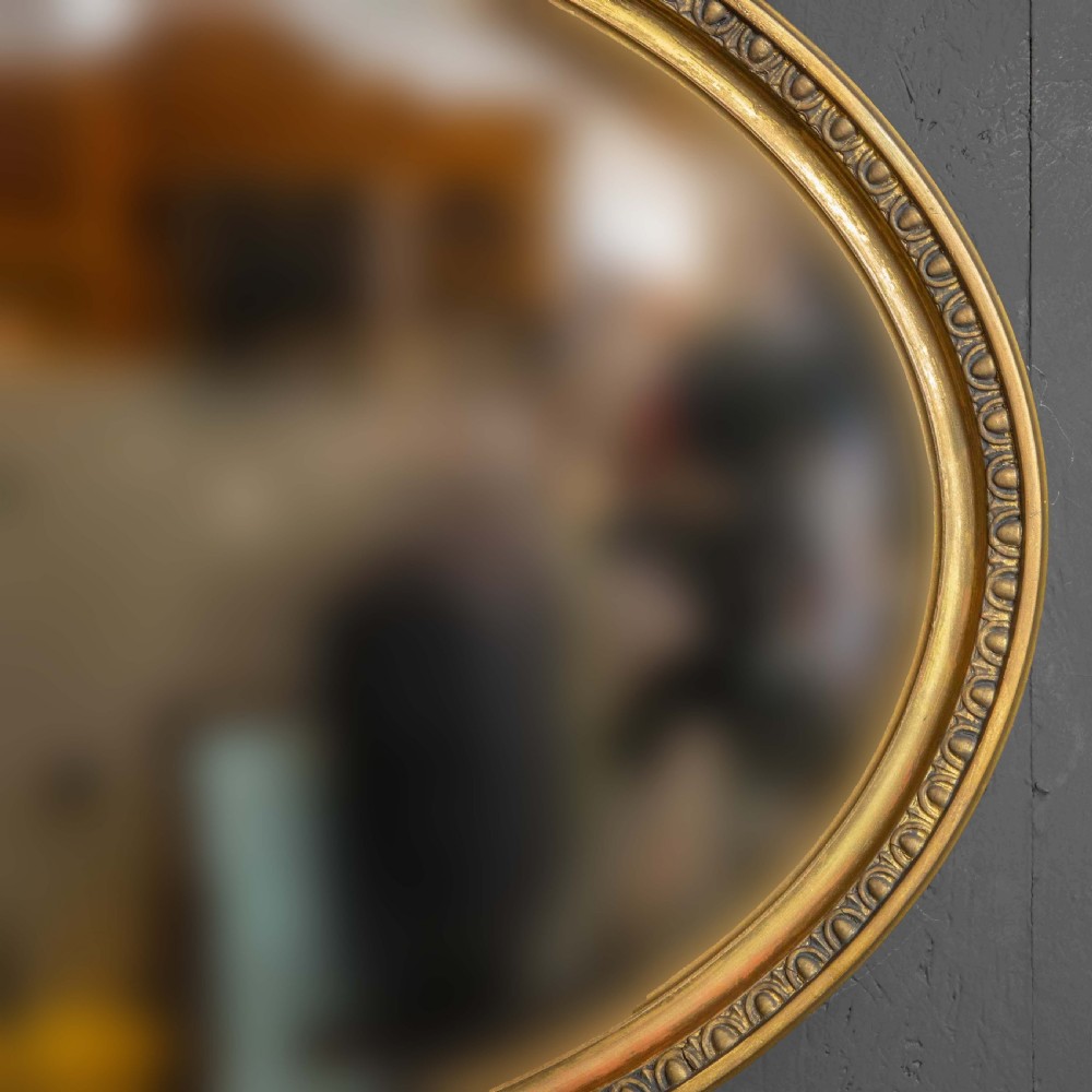 An oval gilded wall mirror decorated with an ornamental urn cresting at the top and floral swags along the lower edge. View V