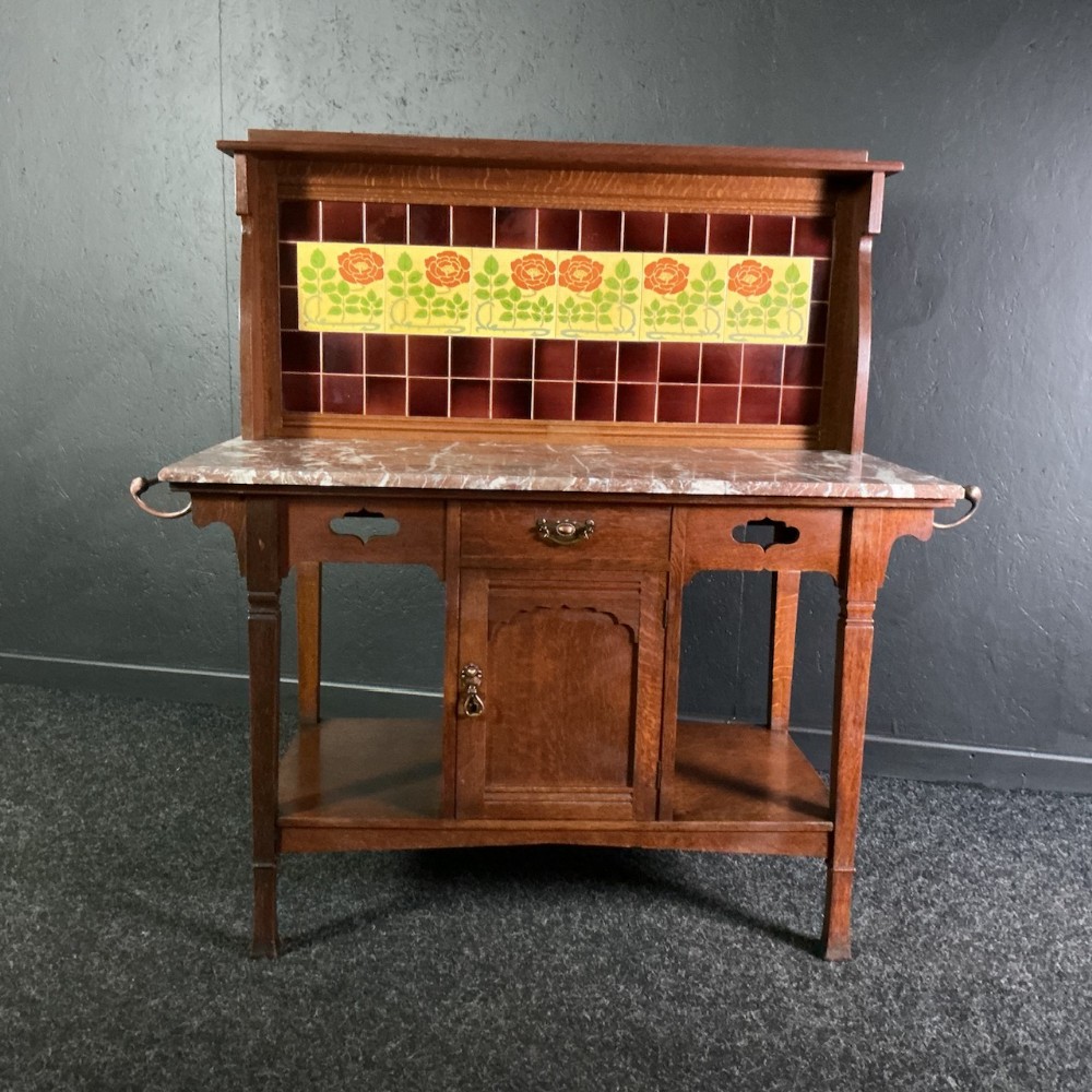 An Arts and Crafts oak washstand featuring a variegated marble top, a tiled splashback with stylized orange roses, and a central cupboard flanked by side towel rails.