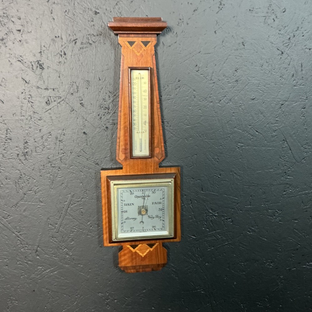 A vertical Art Deco wall-mounted aneroid barometer in warm brown wood with geometric chevron marquetry, an integrated tube thermometer, and a square weather dial.