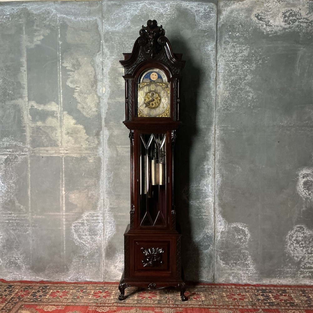 An Edwardian dark mahogany longcase clock featuring a carved scroll pediment, a moonphase dial, a glazed door displaying brass weights, and a carved lower panel.