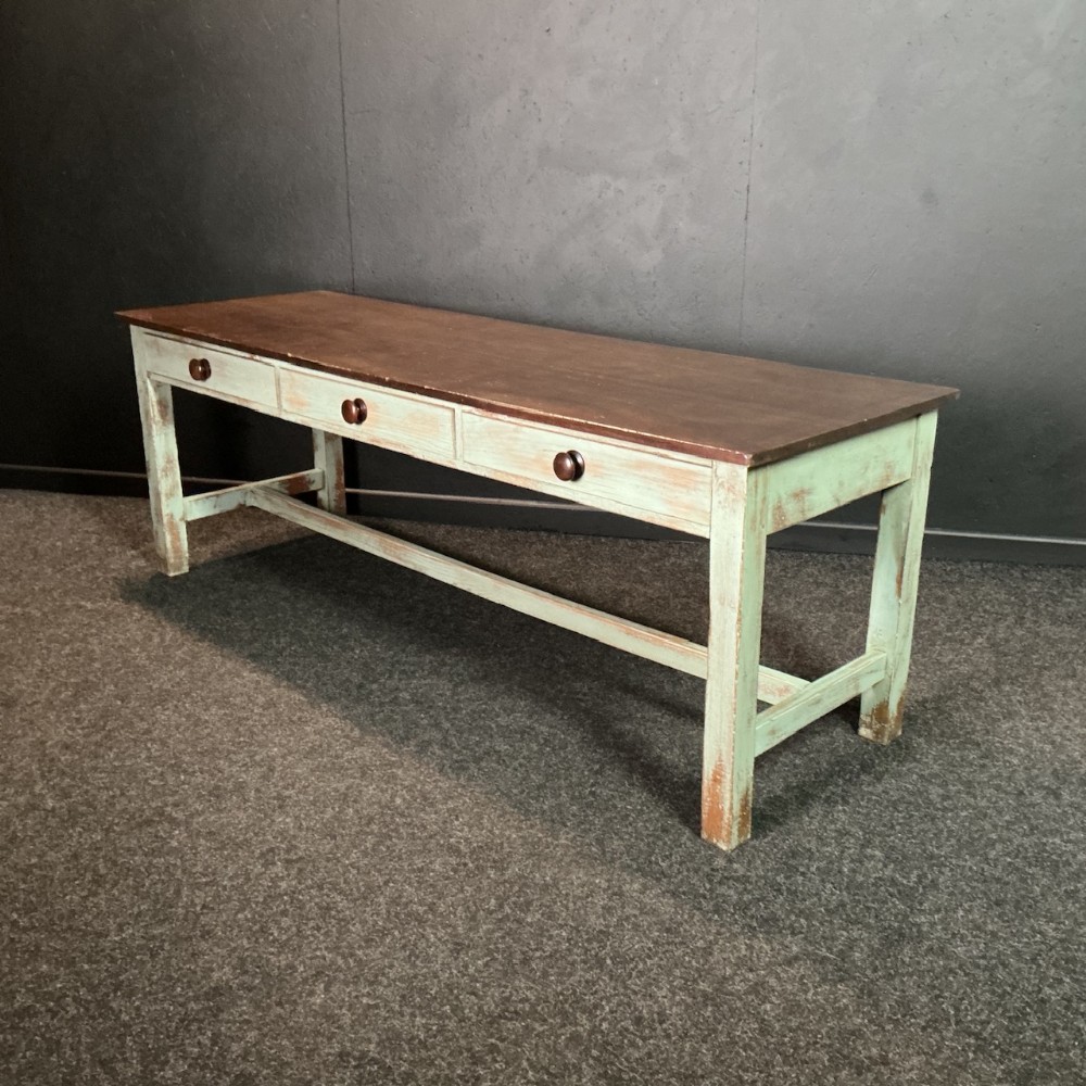A long rectangular antique school dining table featuring a dark wooden top, three frieze drawers with dark turned knobs, an H-stretcher base, and distressed light green paint.
