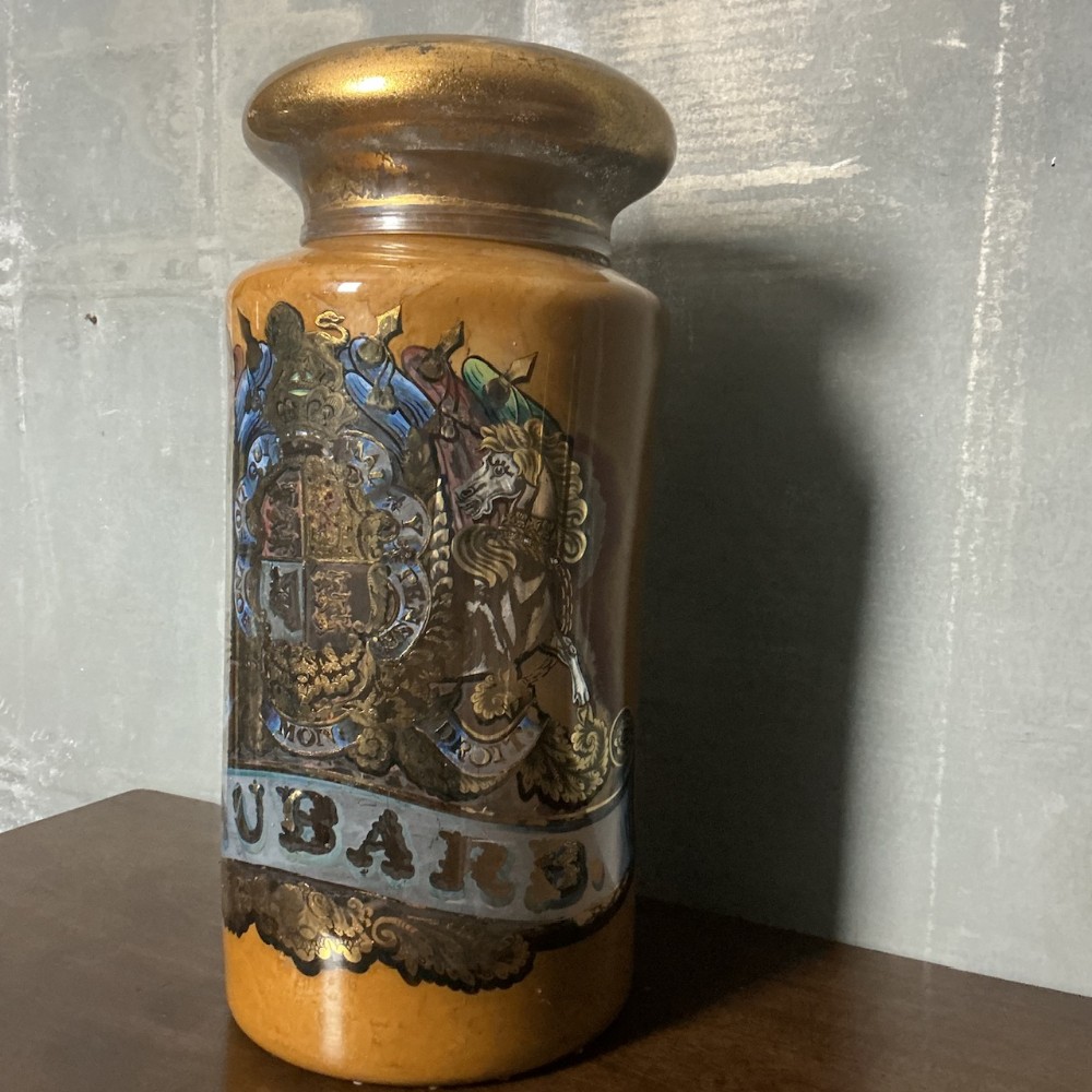 A large Victorian ceramic apothecary jar with an ochre glaze, a gold domed stopper, and hand-painted heraldic artwork above a banner reading RHUBARB.