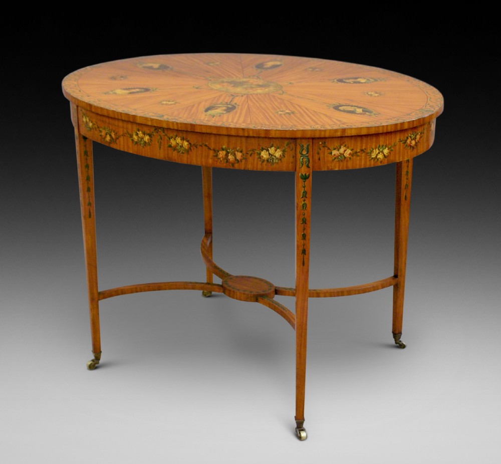 An antique Sheraton Revival oval centre table made of satinwood featuring radiating top veneers, hand-painted floral garlands, tapered legs, and an arched stretcher with brass castors.