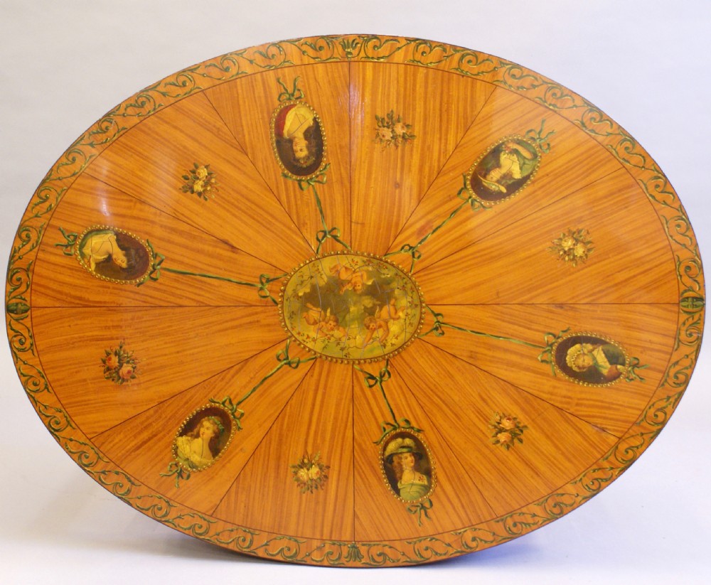 An antique Sheraton Revival oval centre table made of satinwood featuring radiating top veneers, hand-painted floral garlands, tapered legs, and an arched stretcher with brass castors. View II