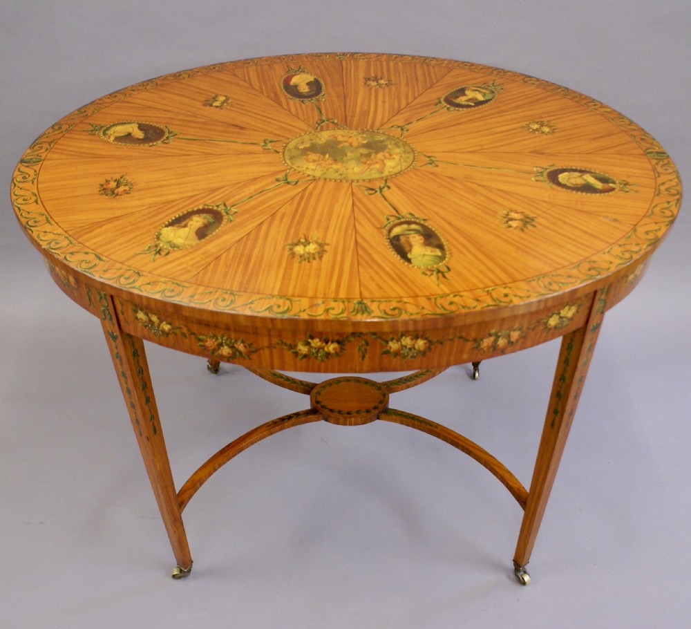 An antique Sheraton Revival oval centre table made of satinwood featuring radiating top veneers, hand-painted floral garlands, tapered legs, and an arched stretcher with brass castors. View V