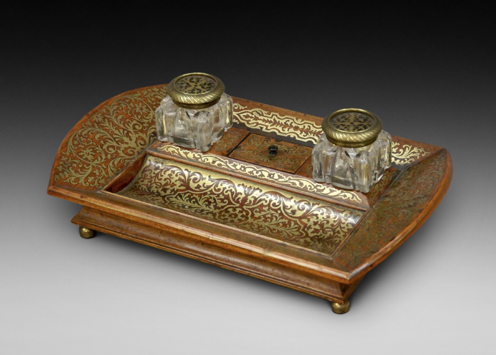 A Regency rosewood desk inkstand decorated with intricate floral brass inlay, featuring a curved pen tray, central lid, two clear cut-glass inkwells with brass caps, and brass ball feet.