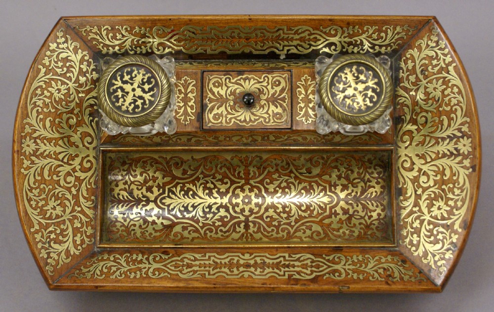 A Regency rosewood desk inkstand decorated with intricate floral brass inlay, featuring a curved pen tray, central lid, two clear cut-glass inkwells with brass caps, and brass ball feet. View II