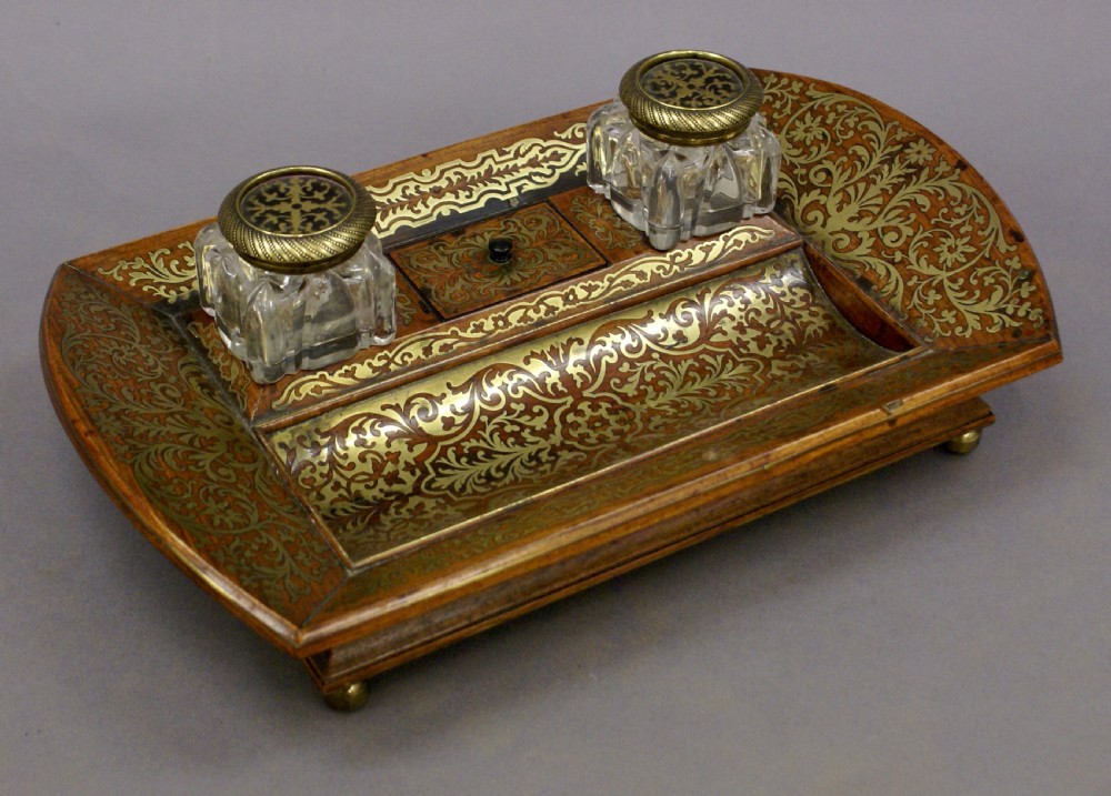 A Regency rosewood desk inkstand decorated with intricate floral brass inlay, featuring a curved pen tray, central lid, two clear cut-glass inkwells with brass caps, and brass ball feet. View V