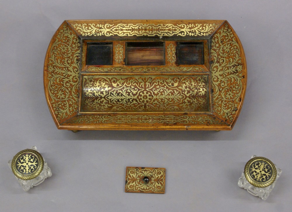 A Regency rosewood desk inkstand decorated with intricate floral brass inlay, featuring a curved pen tray, central lid, two clear cut-glass inkwells with brass caps, and brass ball feet. View VI