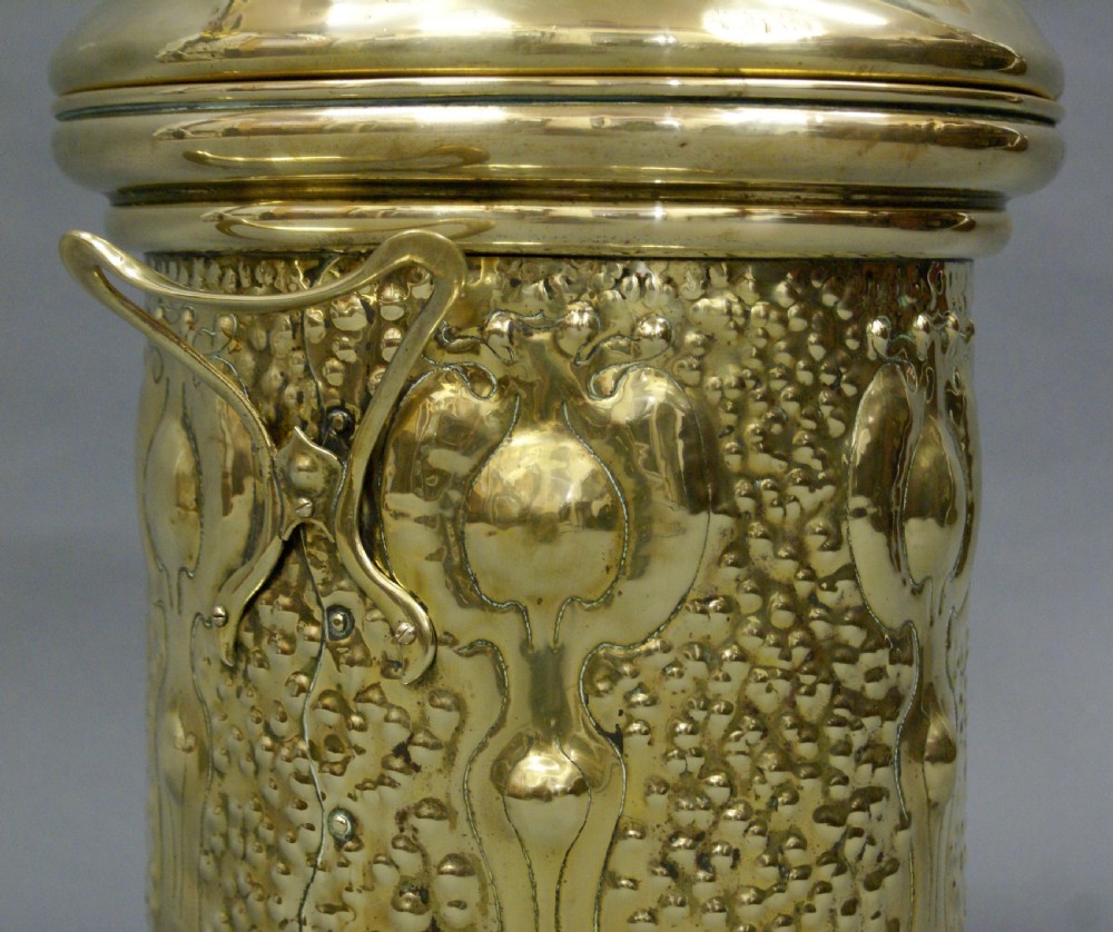 An antique hammered brass cylindrical fireside container with stylized Art Nouveau repousse plant motifs and a domed lid. View II