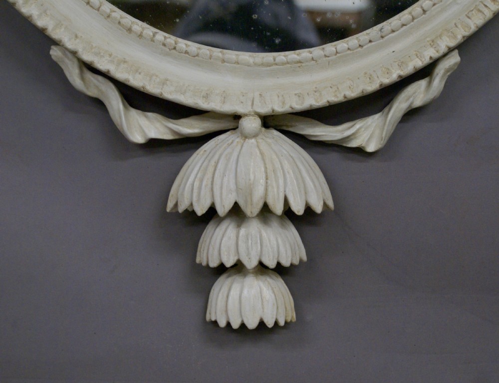 An oval off-white painted Neoclassical wall mirror featuring an urn cresting, delicate floral swags, and decorative drop detail. View III