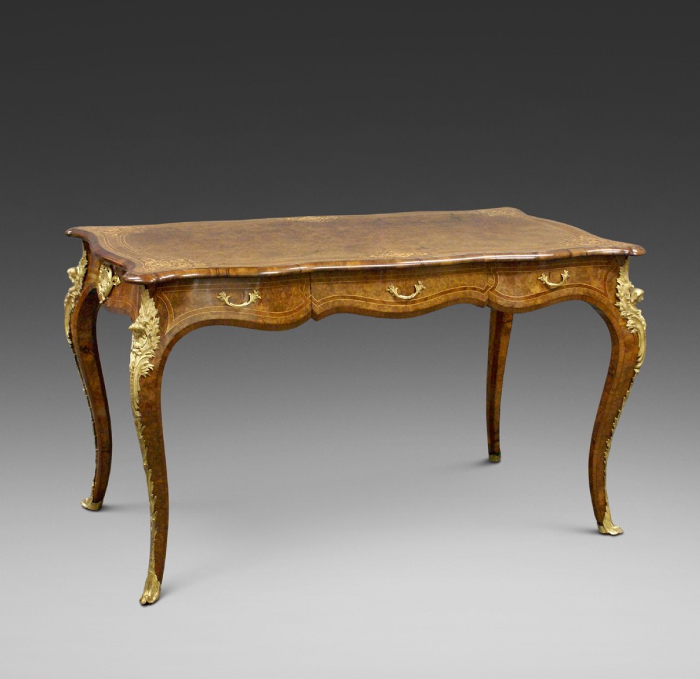 An English burr walnut bureau plat featuring gilt bronze ormolu mounts, a serpentine top, frieze drawers, and cabriole legs.